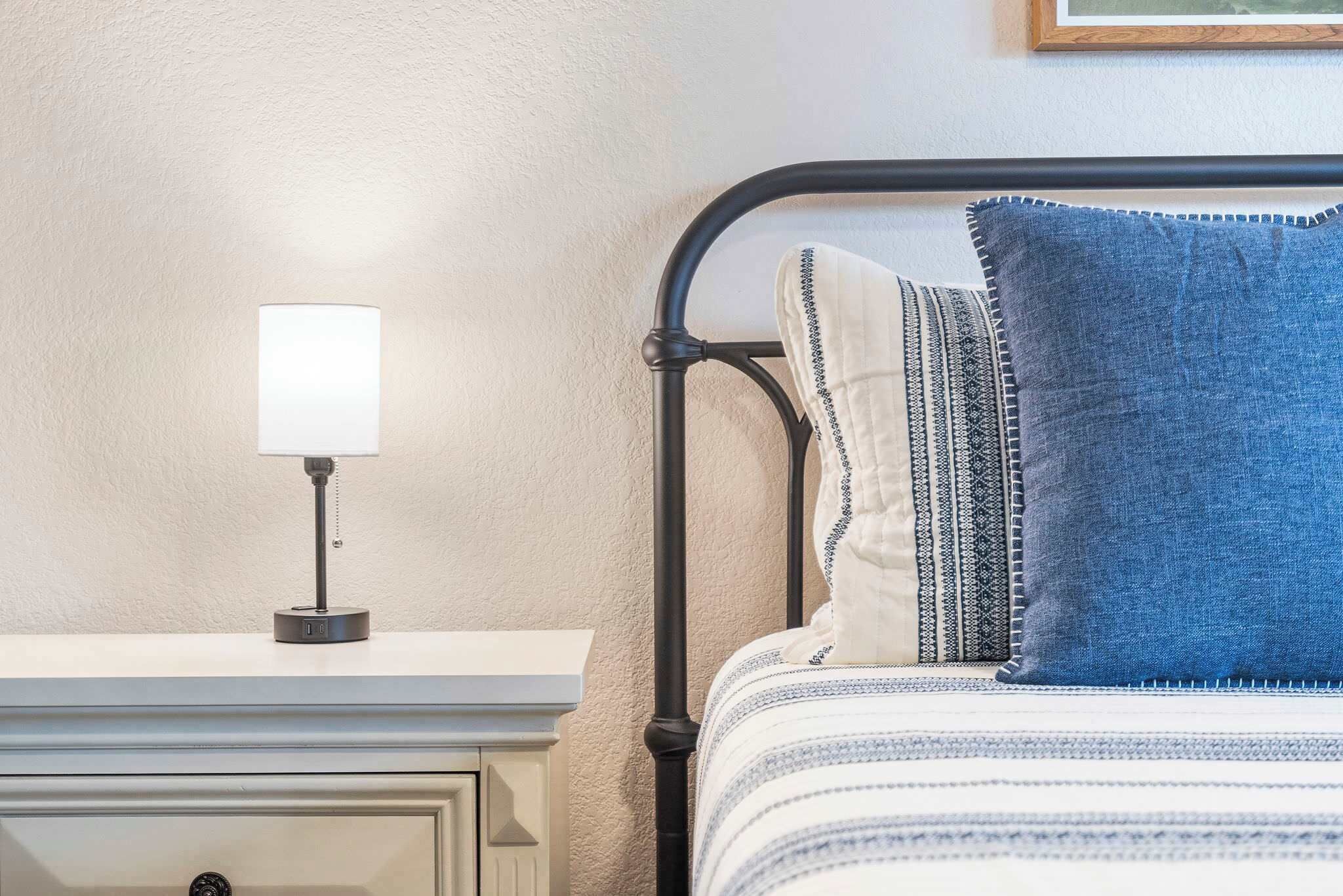 Un lit avec un cadre en métal noir, une literie blanche et un oreiller bleu.