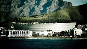 A modern hotel with a unique curved facade, situated by a coastline with a prominent mountain in the background. - Docklands Deluxe Apartments by ITC Hospitality (Cape Town)