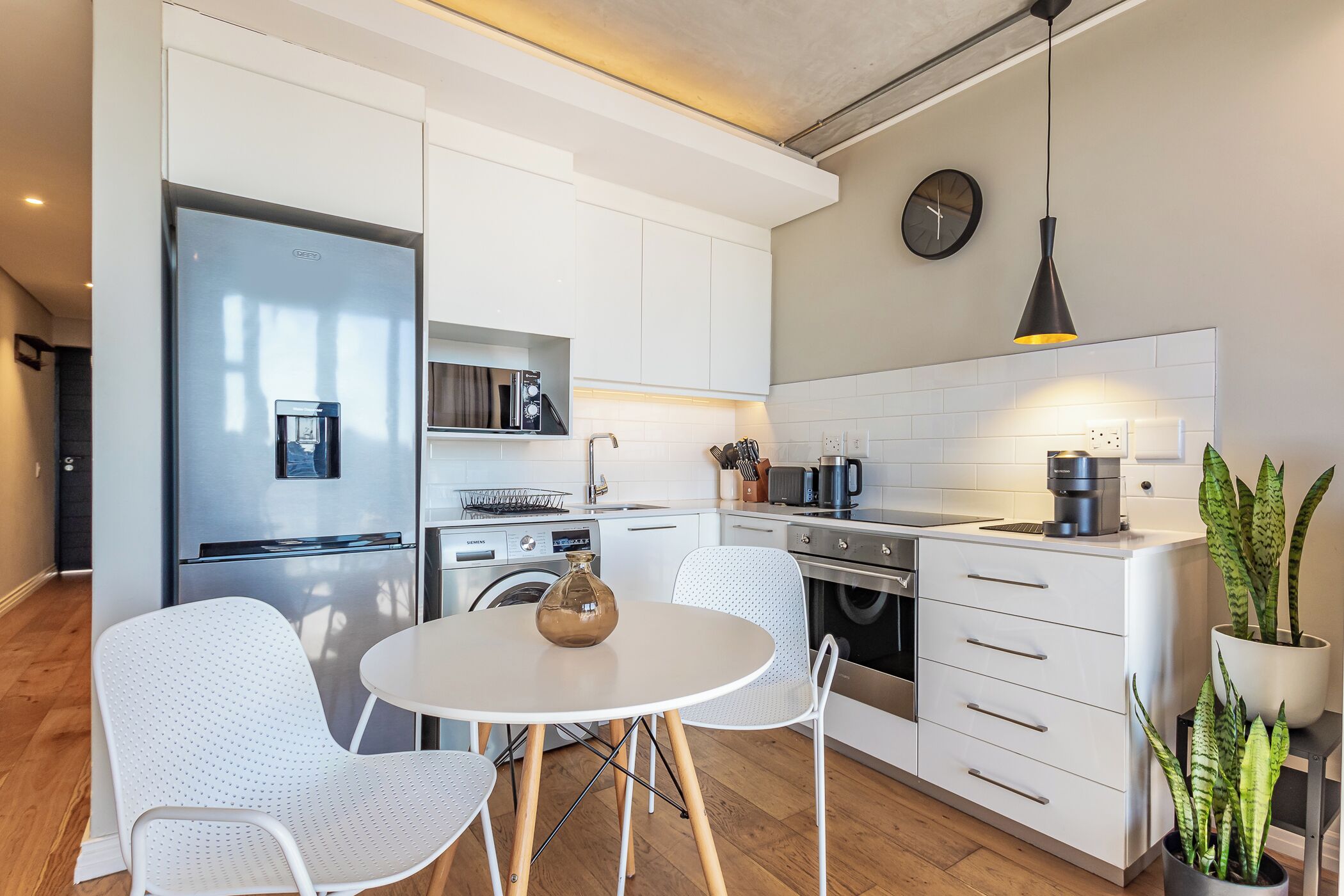 Une cuisine moderne avec des meubles blancs, un réfrigérateur en acier inoxydable, une table de salle à manger ronde avec deux chaises, une machine à café et une horloge au mur.