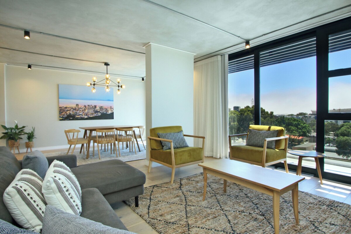 Uma sala de estar moderna com sofá, poltronas, mesa de jantar e uma janela grande com vista para a cidade.