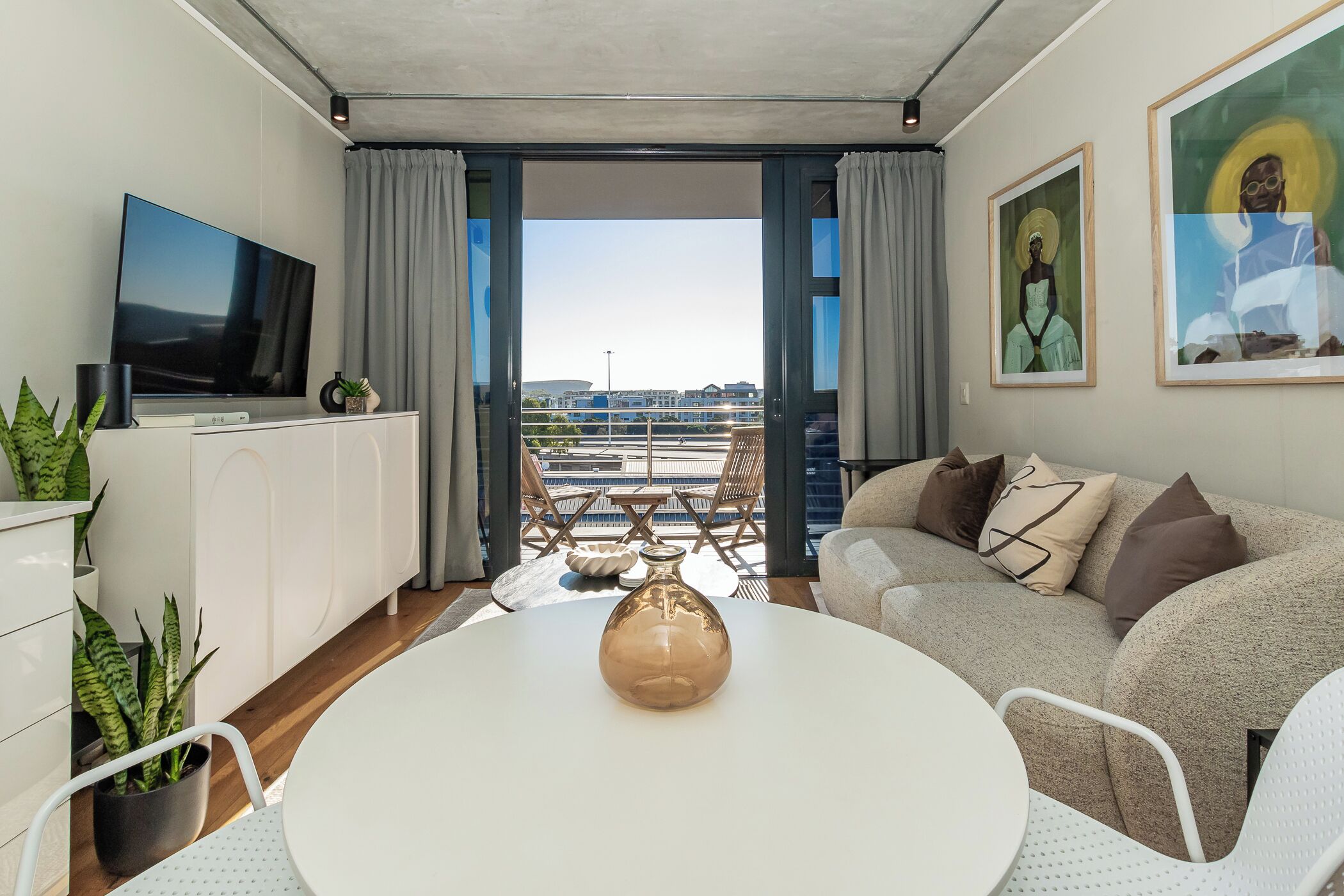 A modern living room with a flat-screen TV, a white dining table with a vase, a sofa with cushions, and a balcony with a view.
