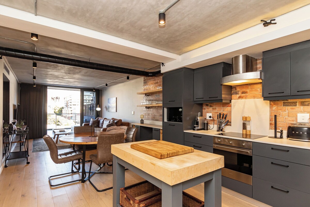 Uma cozinha moderna com armários escuros, uma ilha central e um espaço de jantar com mesa e cadeiras de madeira.