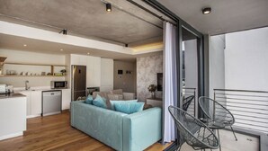 A modern apartment with a living area featuring a light blue sofa, a kitchen with white cabinets, and a balcony with metal chairs. - Docklands Deluxe Apartments by ITC Hospitality (Cape Town)