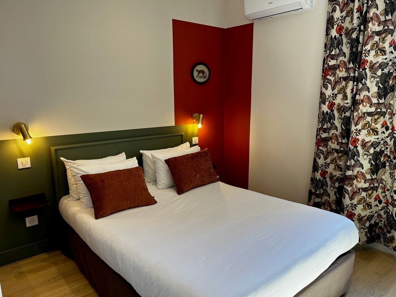 Un lit bien fait, avec des oreillers blancs et bruns, un mur d’accent rouge et un luminaire fixé au mur.