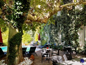 An outdoor patio with tables and chairs, surrounded by lush greenery and a tree. - Cit'Hotel le Louvre (Orange)