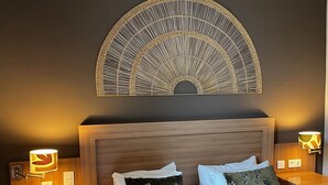 Une chambre d’hôtel avec une tête de lit en bois, deux tables de chevet avec des lampes, un lit avec des oreillers blancs et à motifs, et un élément décoratif mural au-dessus du lit.