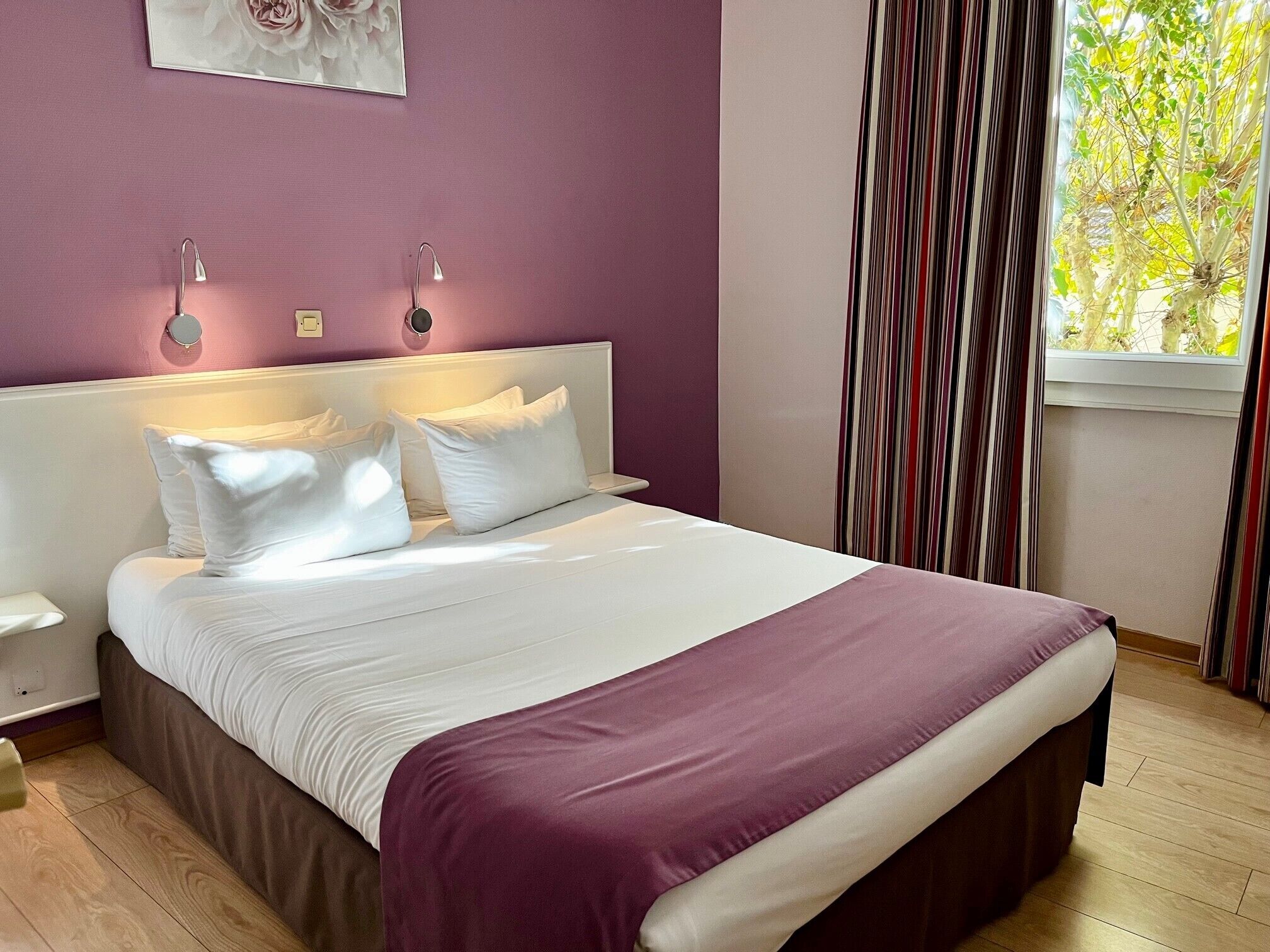 A bed with white linens and a purple throw, a nightstand, a wall clock, and a window with curtains.