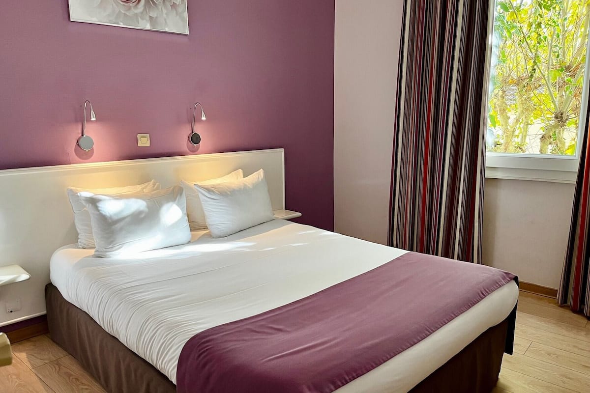A bed with white linens and a purple throw, a nightstand, a wall clock, and a window with curtains.