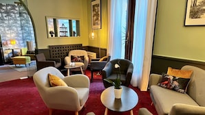 Un salon moderne avec un tapis à motifs rouges, deux fauteuils, un canapé, une petite table ronde ornée d’un pot de plantes et une fenêtre donnant sur des arbres.