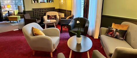 A modern living room with a red patterned carpet, two armchairs, a sofa, a small round table with a potted plant, and a window with a view of trees.