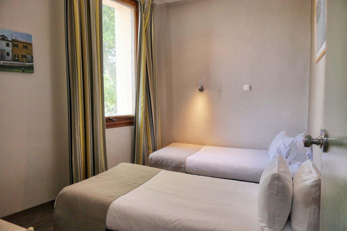 A room with two beds, a window with curtains, a wall-mounted light, and a door with a handle.