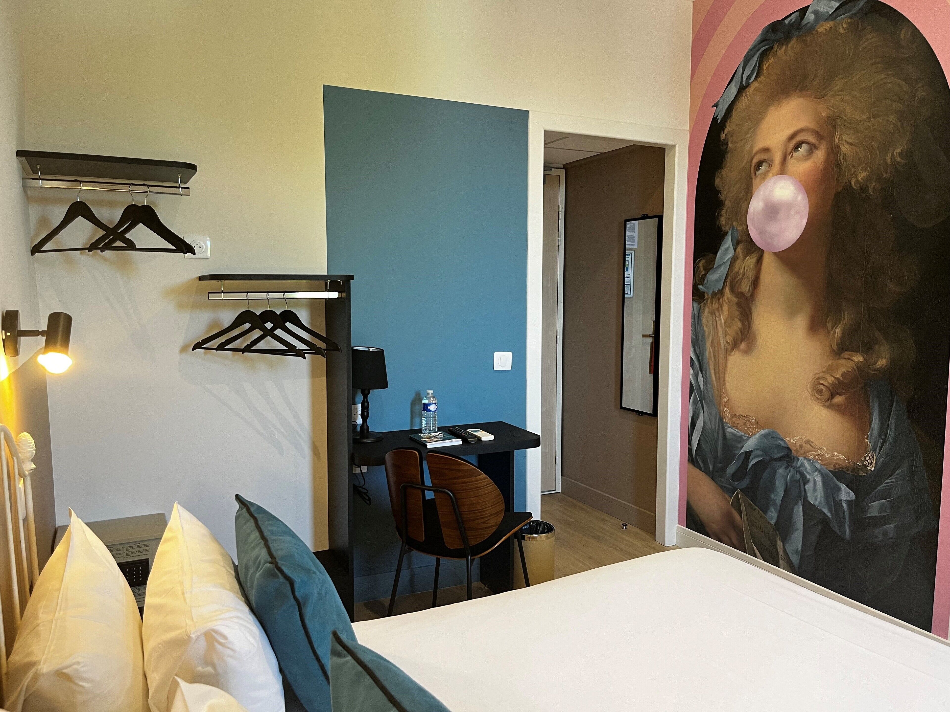 A hotel room with a bed, a desk, a chair, a water bottle, and a painting of a woman on the wall.