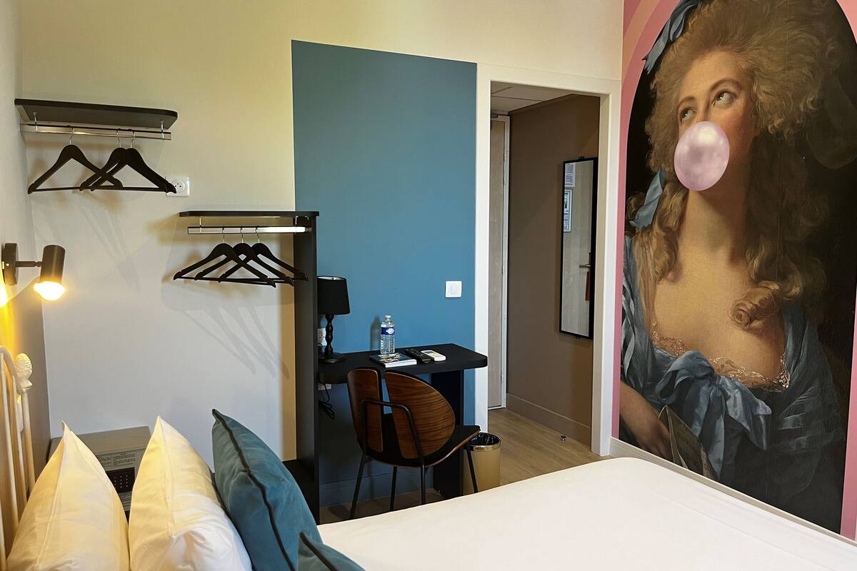 A hotel room with a bed, a desk, a chair, a water bottle, and a painting of a woman on the wall.