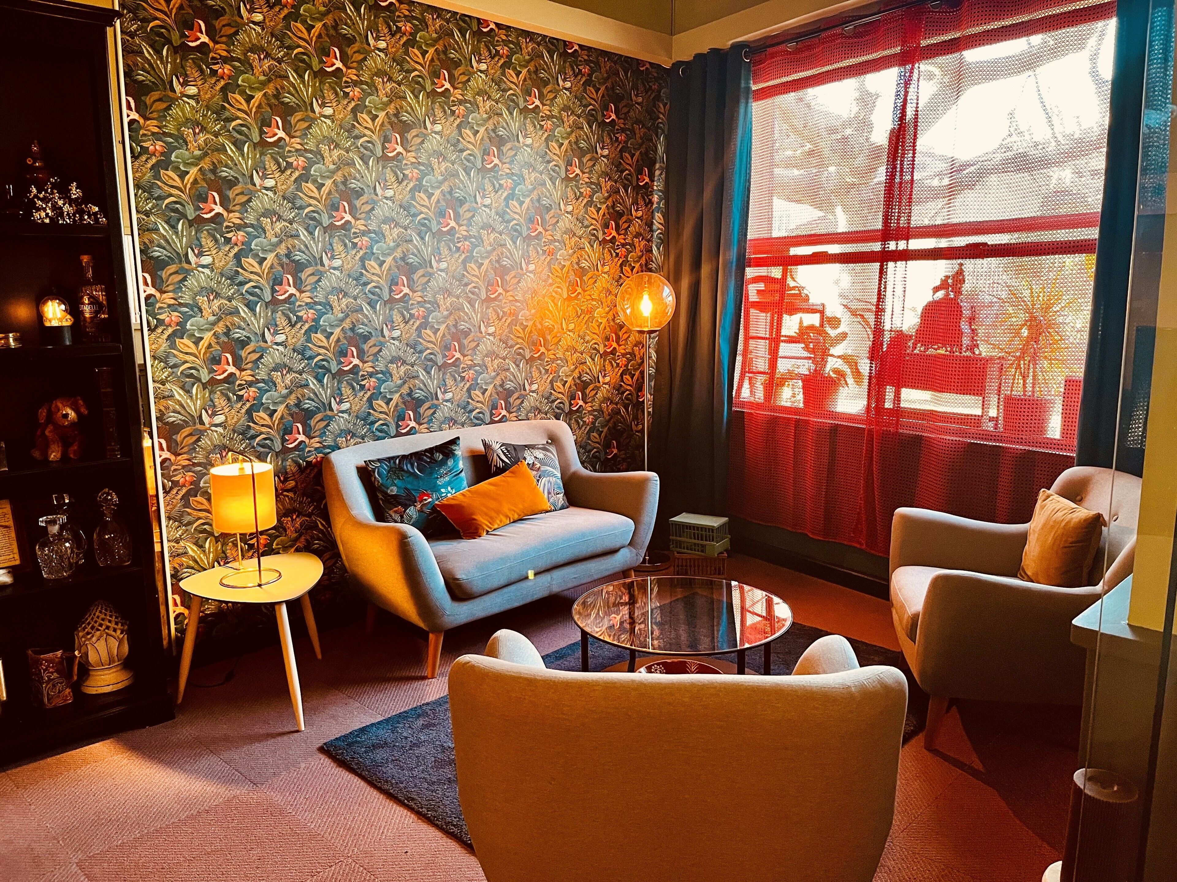 Uma sala de estar acolhedora com papel de parede estampado, um sofá, poltronas, uma mesa de centro de vidro e uma luminária.