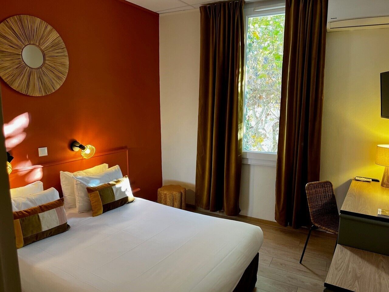 Una habitación de hotel con una cama grande, un escritorio con lámpara, una silla, un espejo redondo en la pared y una ventana con cortinas.