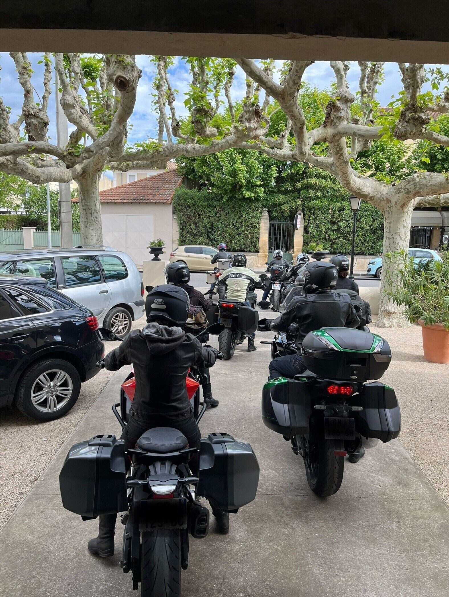 A group of motorcyclists parked in a covered area with trees and parked cars in the background.
