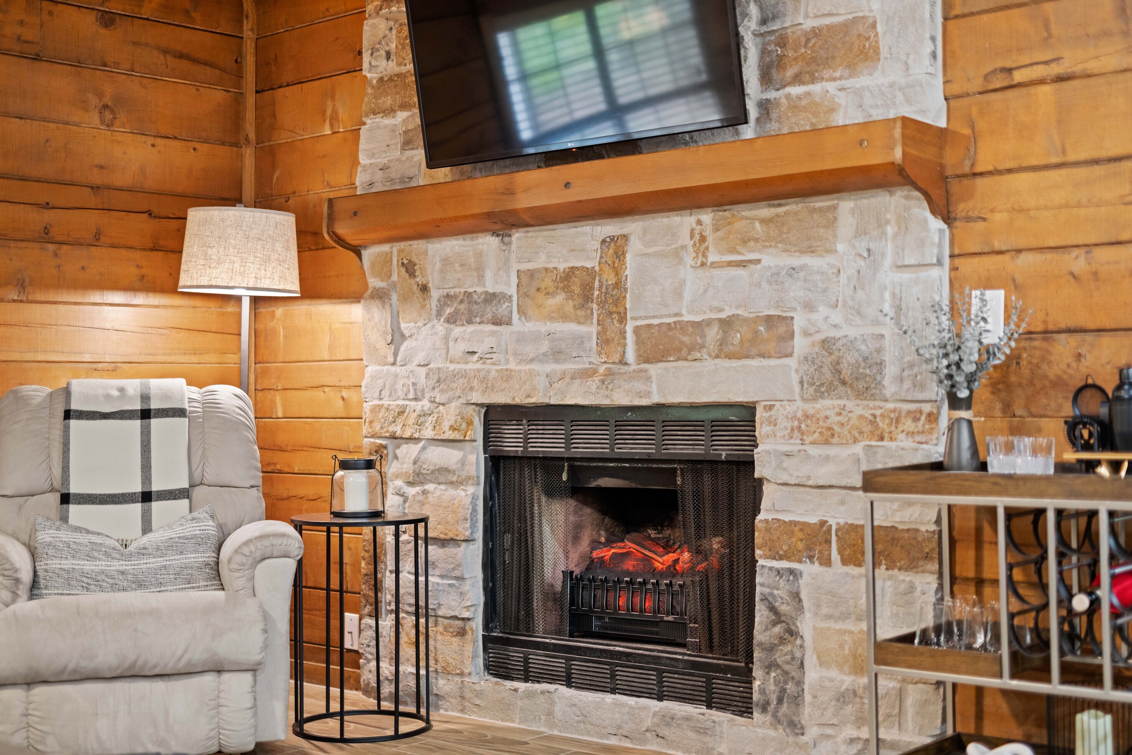 A cozy living room with a stone fireplace, a comfortable armchair, a TV mounted above the fireplace, and a small side table with a lamp.