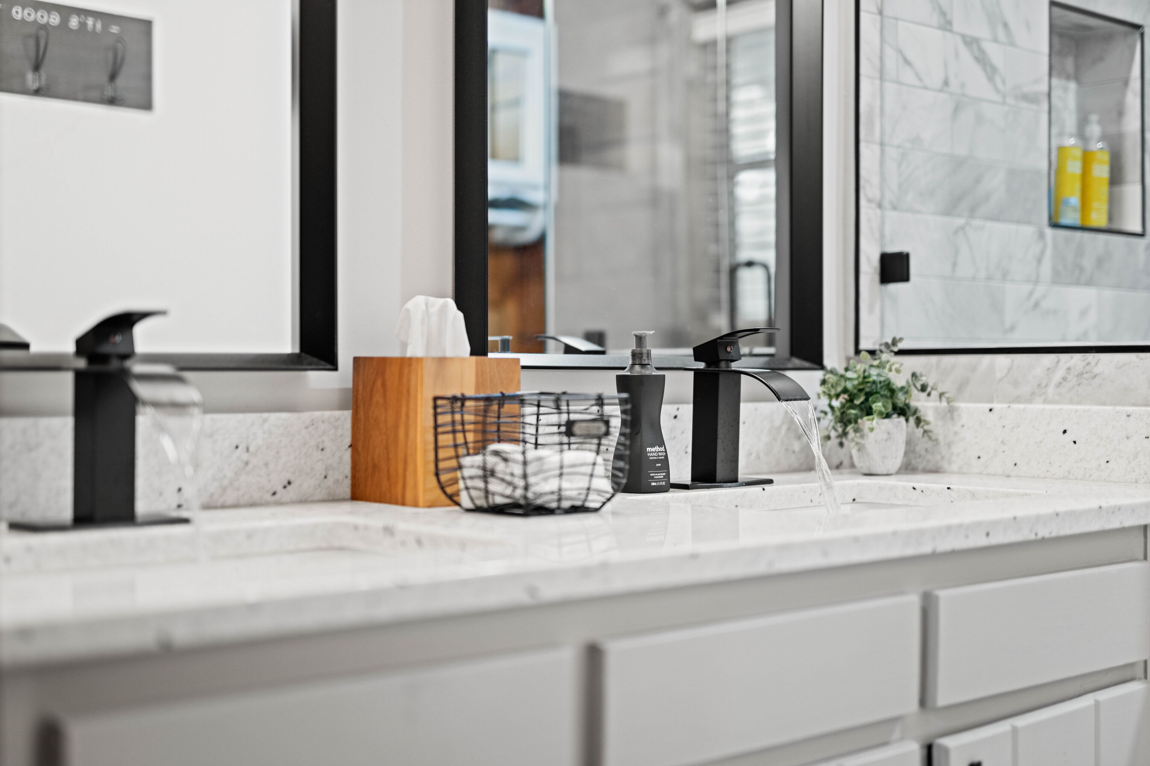 Un baño moderno con encimera de mármol, tabla de madera, dispensador de jabón negro y una planta en maceta.