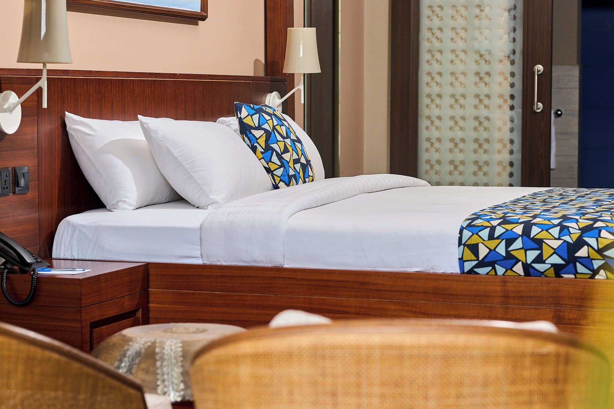 A neatly made bed with white bedding and a colorful geometric-patterned throw. There is a wooden nightstand with a telephone and a lamp.