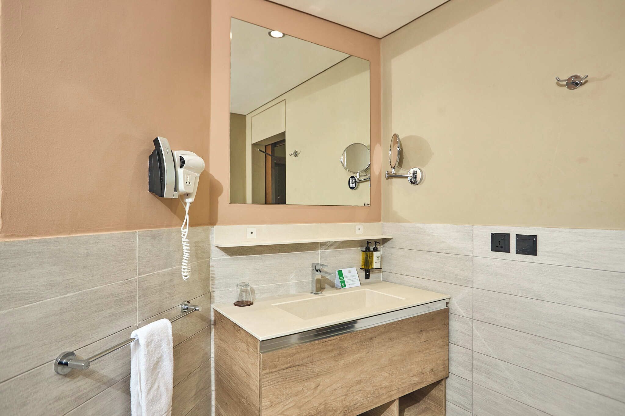 A modern bathroom with a beige vanity, a large mirror, a hairdryer, and a towel rack.