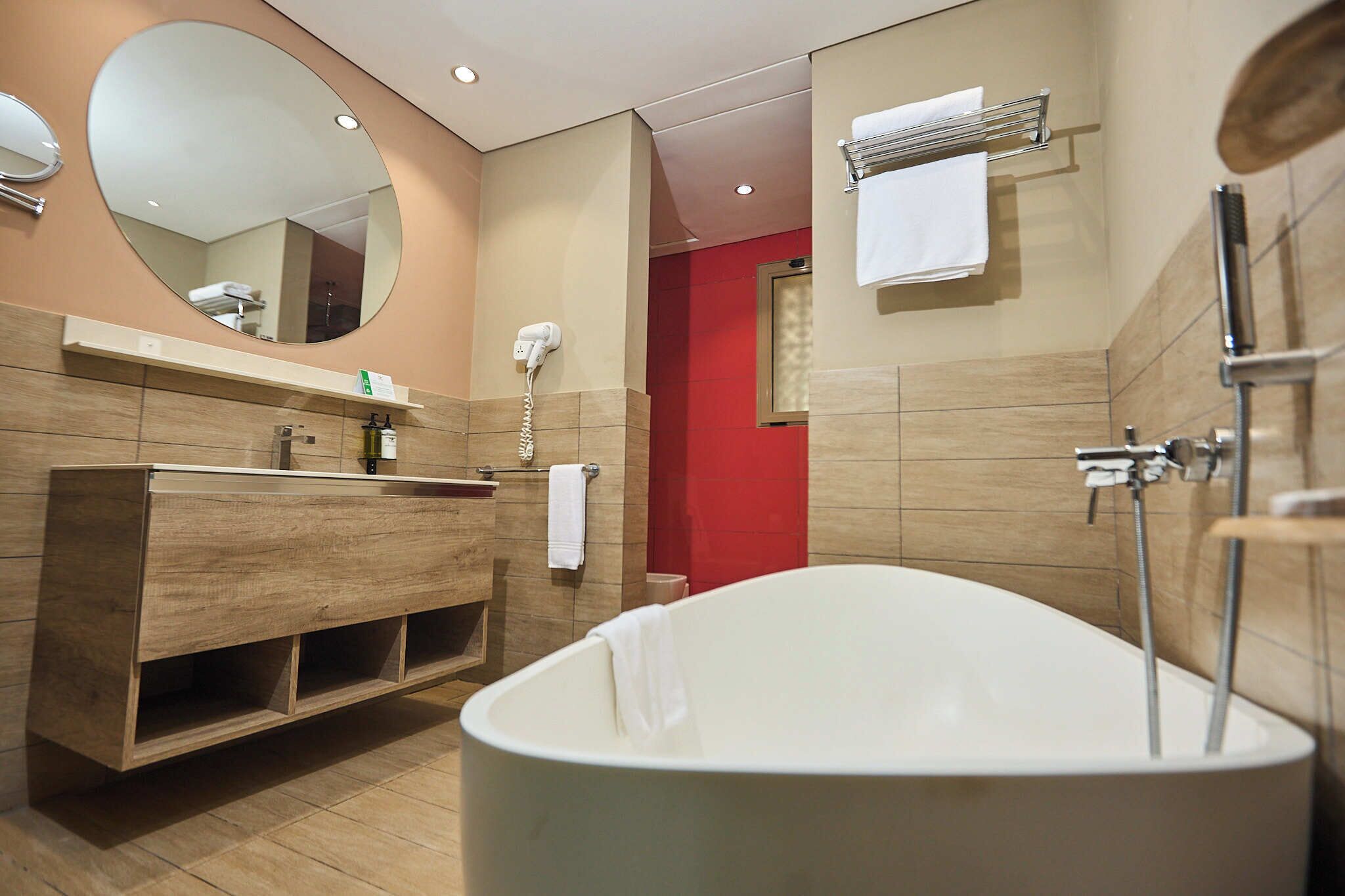 A modern bathroom with a freestanding bathtub, a wooden vanity, a large mirror, and a red accent wall.