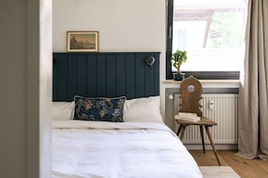 Un dormitorio con cama, una silla, un radiador, una ventana con cortinas y un cuadro en la pared.