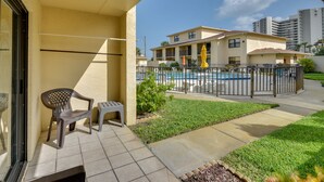 A balcony with a chair and a small table, a pool area with a fence, and a building in the background. - Daytona Beach Shores Condo Rental: Walk to Beach! (Daytona Beach Shores)