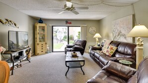 A living room with a brown leather sofa, a coffee table, a TV, a ceiling fan, and a view of the outdoors through a glass door. - Daytona Beach Shores Condo Rental: Walk to Beach! (Daytona Beach Shores)