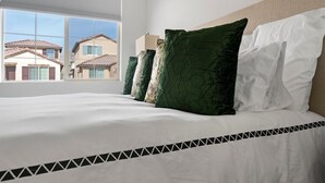 Ein ordentlich bezogenes Bett mit weißen Leinen und einer schwarzen geometrischen Kante, mit grünen und weißen Kissen, und ein Fenster mit Blick auf Häuser.