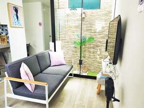 A modern living room with a grey sofa, a flat-screen TV, and a glass door leading to a balcony with a plant.
