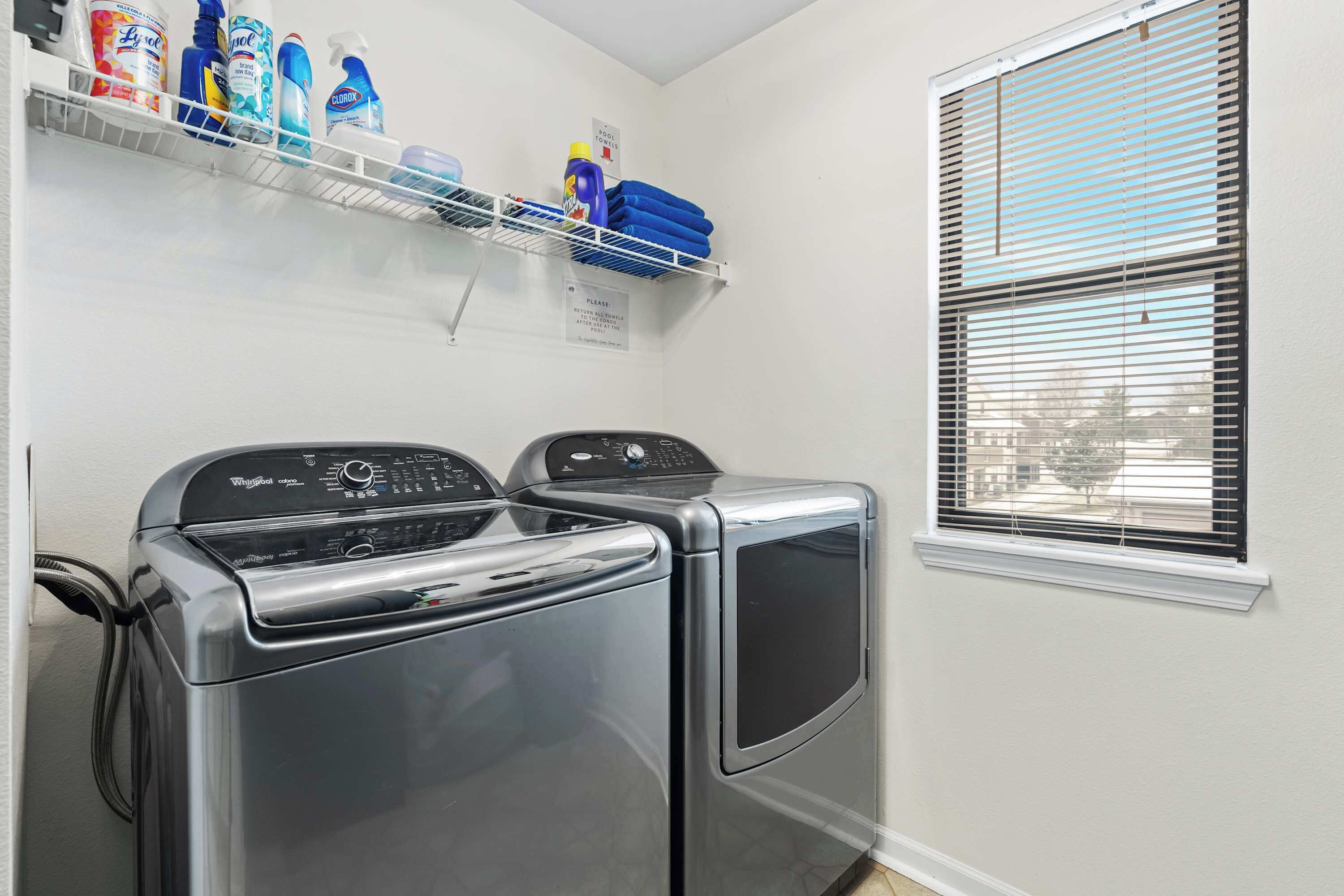 A laundry area with two front-loading washers, a shelf with cleaning supplies, and a window with blinds.
