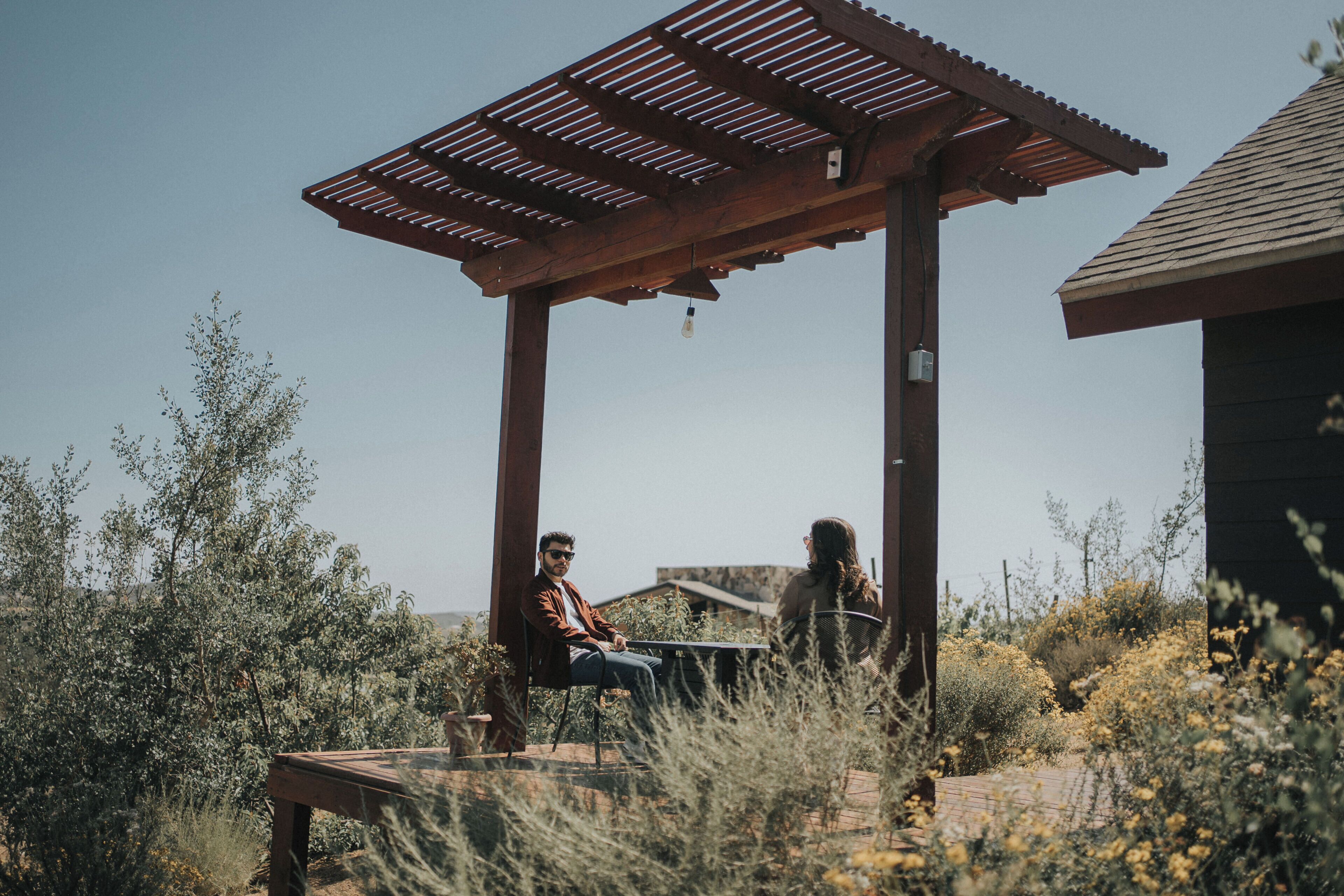 Una pérgola de madera con una mesa y sillas, rodeada de vegetación desértica.
