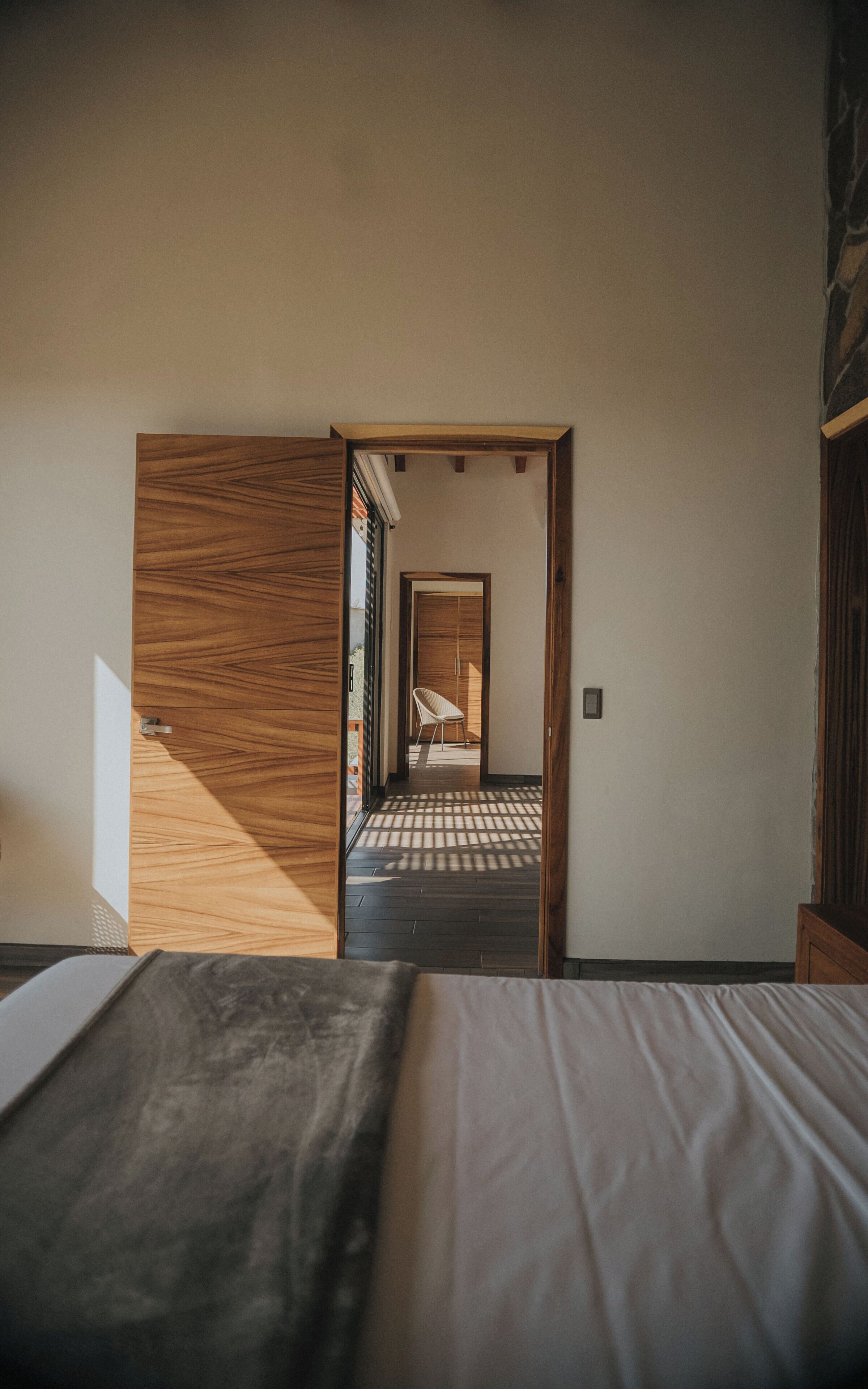 A bedroom with a bed, a wooden wardrobe, and a view of a hallway with a chair.