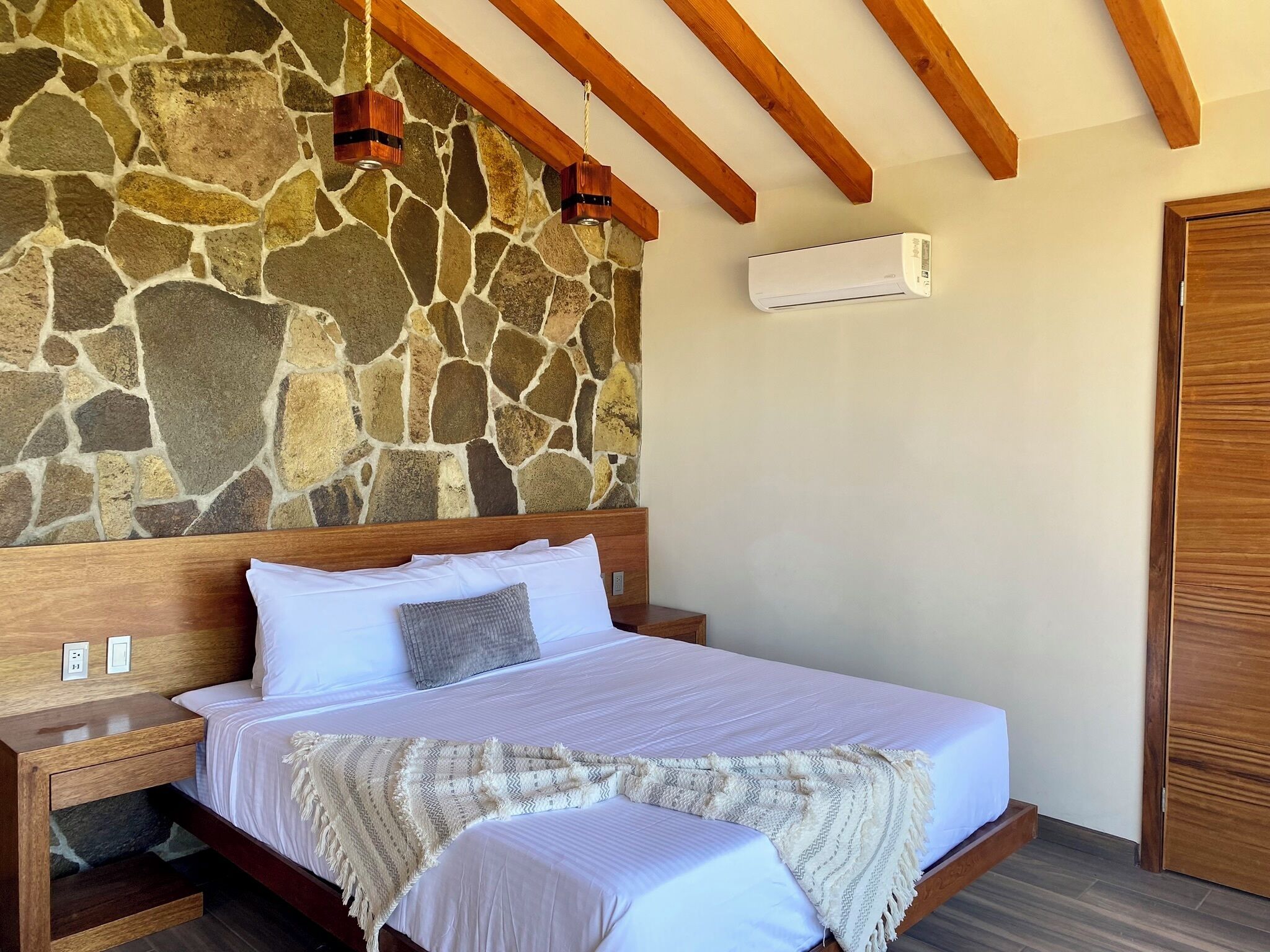 A bedroom with a stone wall, wooden beams, a bed with white bedding, a bedside table, and a wardrobe.