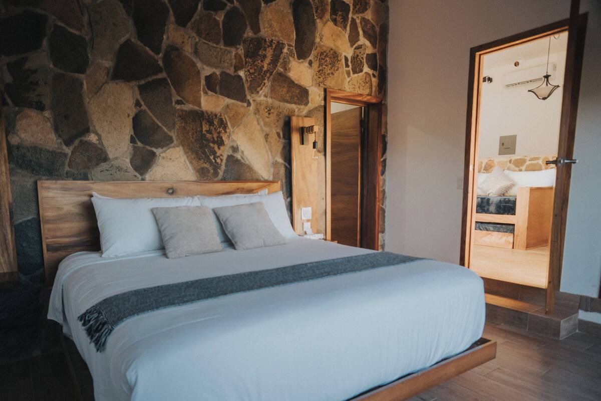 Un dormitorio con pared de piedra, estructura de cama de madera y vistas a un baño por una puerta abierta.