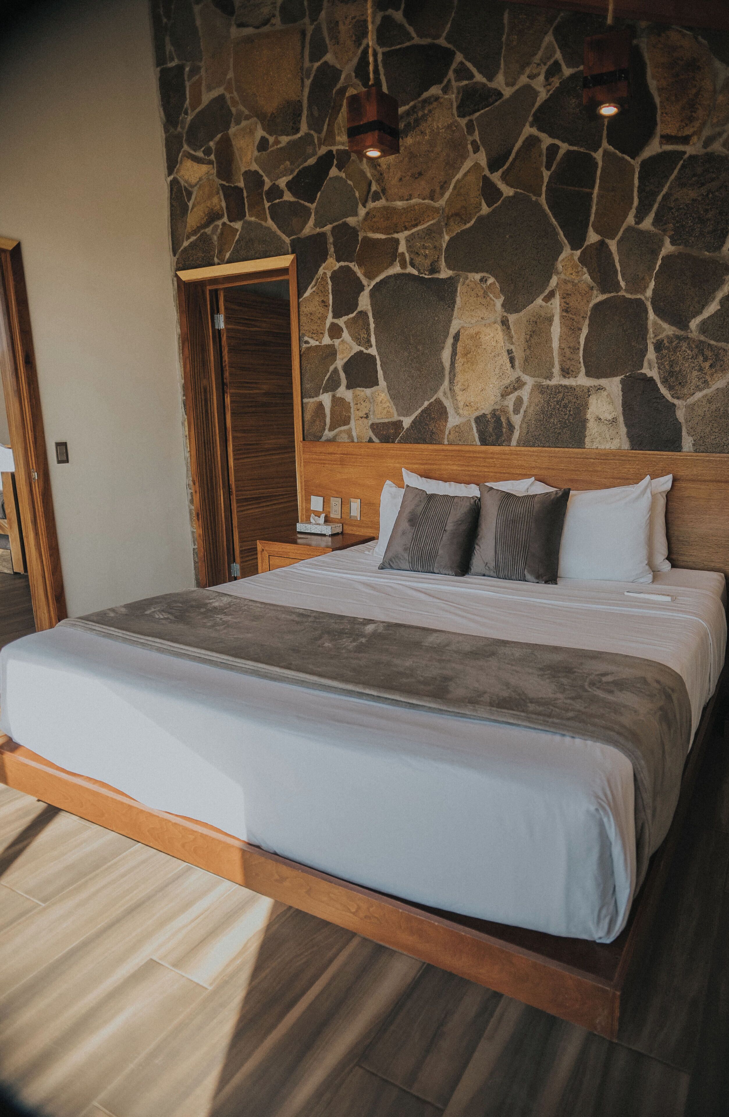 Un dormitorio con pared de piedra, cabecero de madera y cama con colcha y almohadas grises.