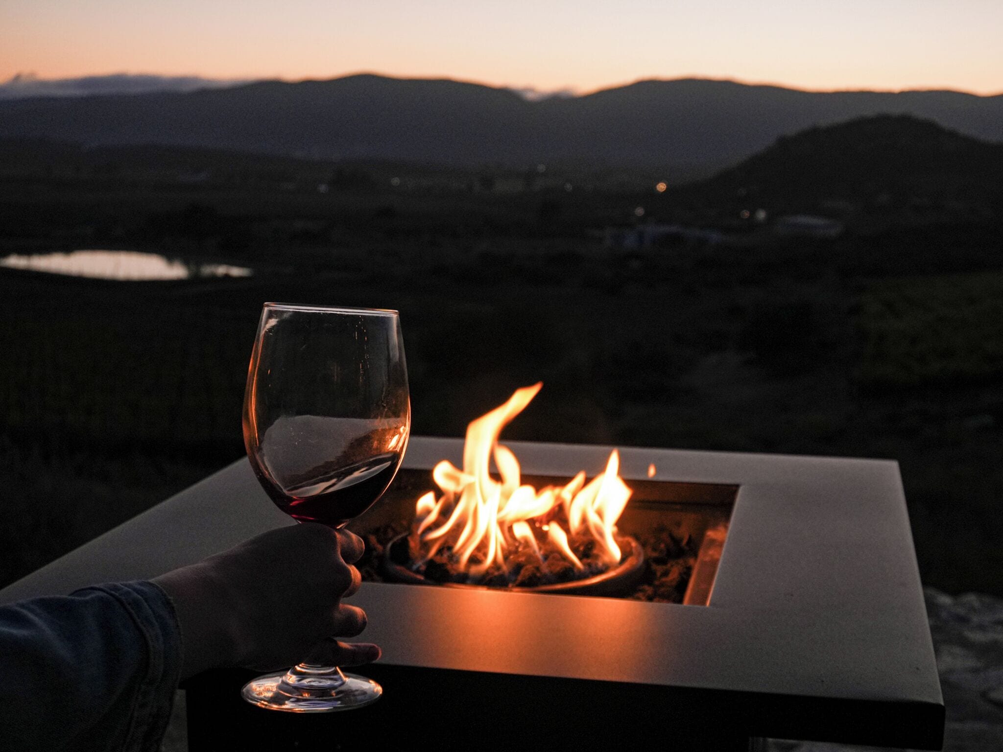 A hand holding a wine glass over a fire pit with a view of mountains and a lake in the background.