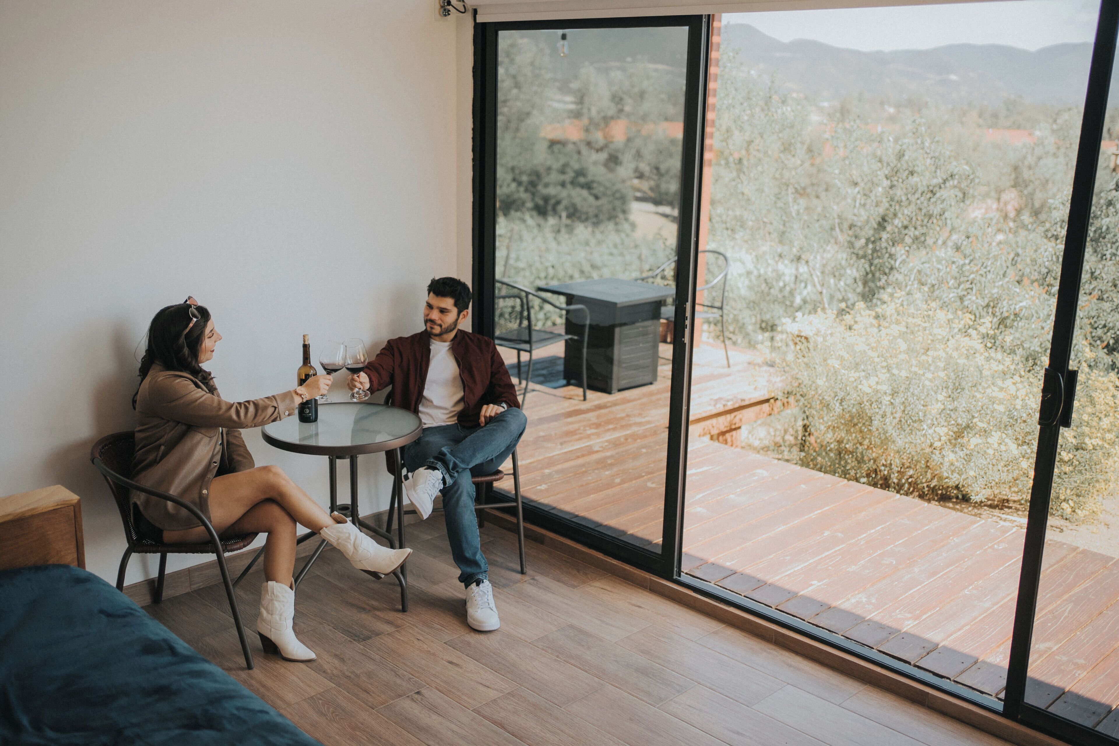 Una pareja sentada a una mesa con una botella de vino, una copa y un menú. La habitación tiene un gran ventanal corredero que da a una terraza exterior con vistas a árboles y montañas.