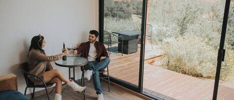 Una pareja sentada a una mesa con una botella de vino, una copa y un menú. La habitación tiene un gran ventanal corredero que da a una terraza exterior con vistas a árboles y montañas.