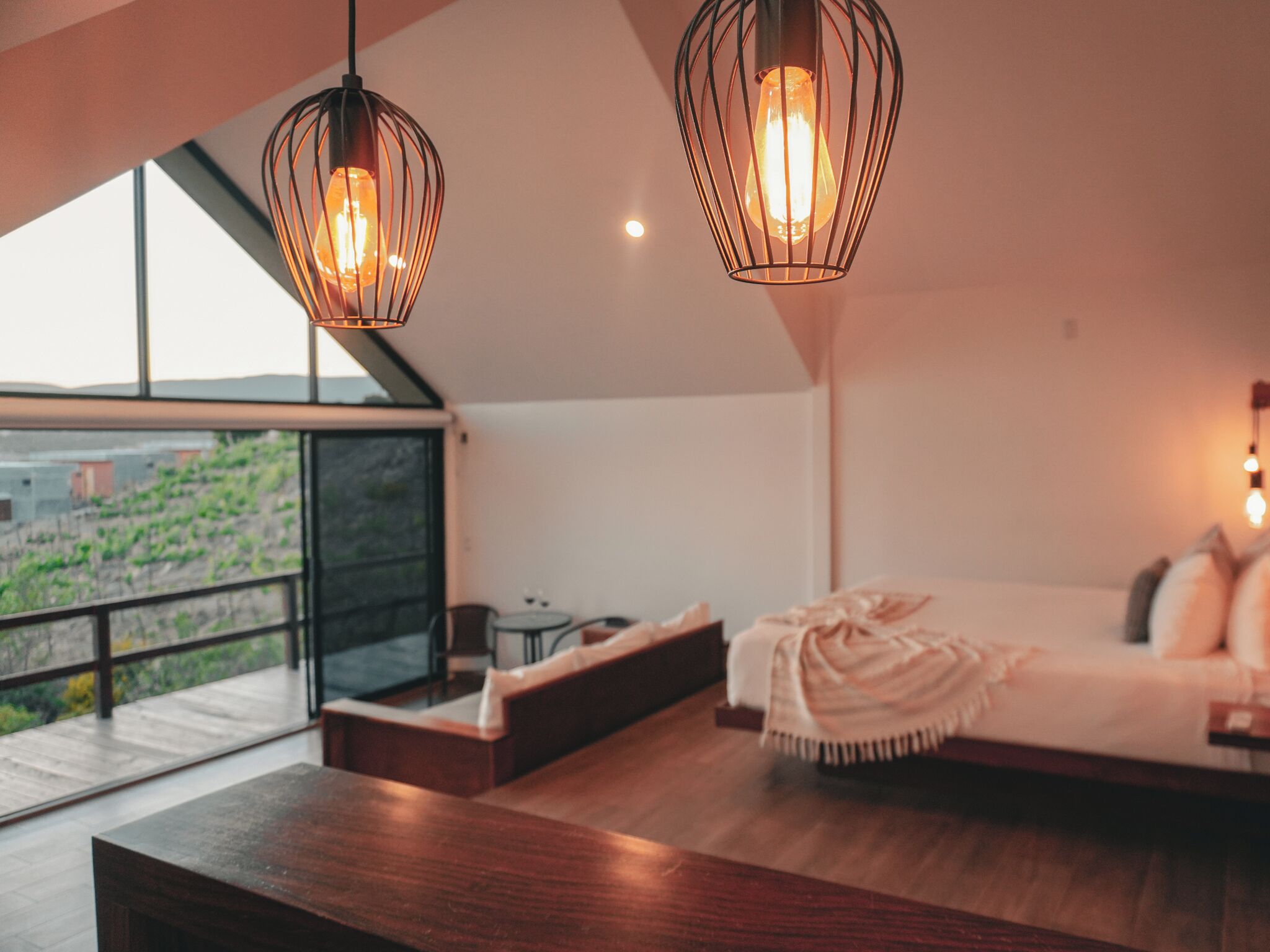 Un intérieur moderne avec une grande fenêtre, un banc en bois, un canapé avec une couverture et deux luminaires suspendus.