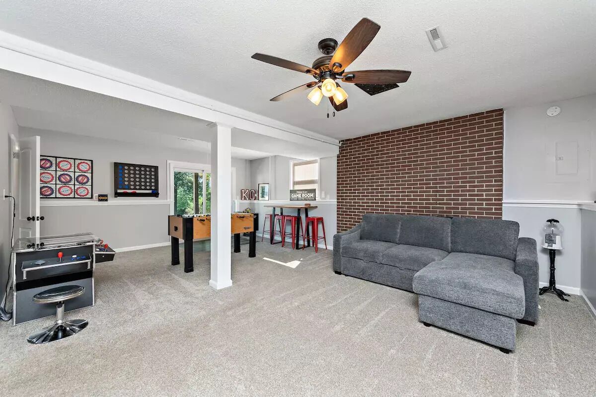 A living room with a gray sofa, a ceiling fan, a brick wall, and a foosball table.