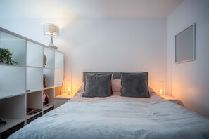 Ein ordentlich eingerichtetes Schlafzimmer mit einem Bett, zwei Nachttischlampen, einer weißen Regaleinrichtung mit Dekoration und einem gerahmten Bild an der Wand.