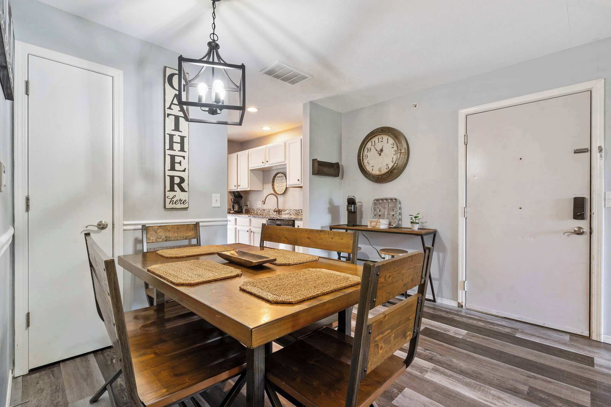 A dining area with a wooden table and chairs, a hanging light fixture, a clock on the wall, and a kitchen in the background.