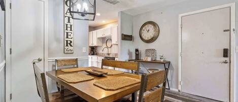 A dining area with a wooden table and chairs, a hanging light fixture, a clock on the wall, and a kitchen in the background.