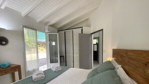 Um quarto com cama, cabeceira de madeira, mesa de cabeceira, janela com cortinas e porta que dá para outro cômodo.