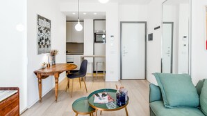 Una sala moderna con una mesa de comedor de madera, una mesa de centro verde y un sofá color turquesa. Se observa una cocina al fondo, con un microondas y armarios.