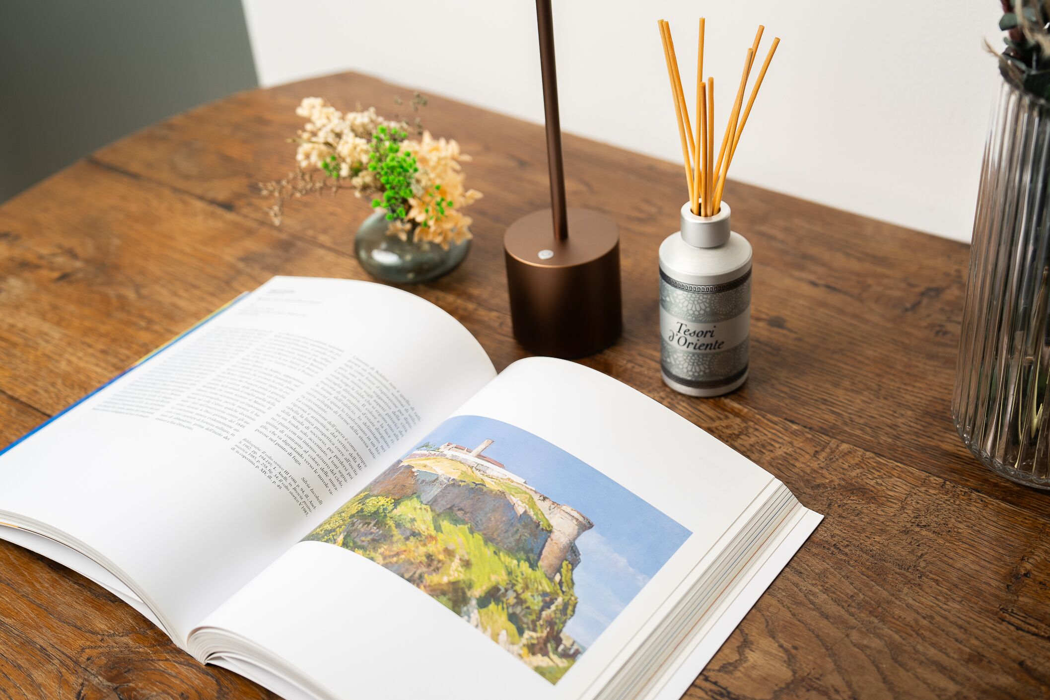 Un libro aperto con una fotografia sulla pagina destra, un vaso con canne palustri e una piccola pianta su un tavolo di legno.