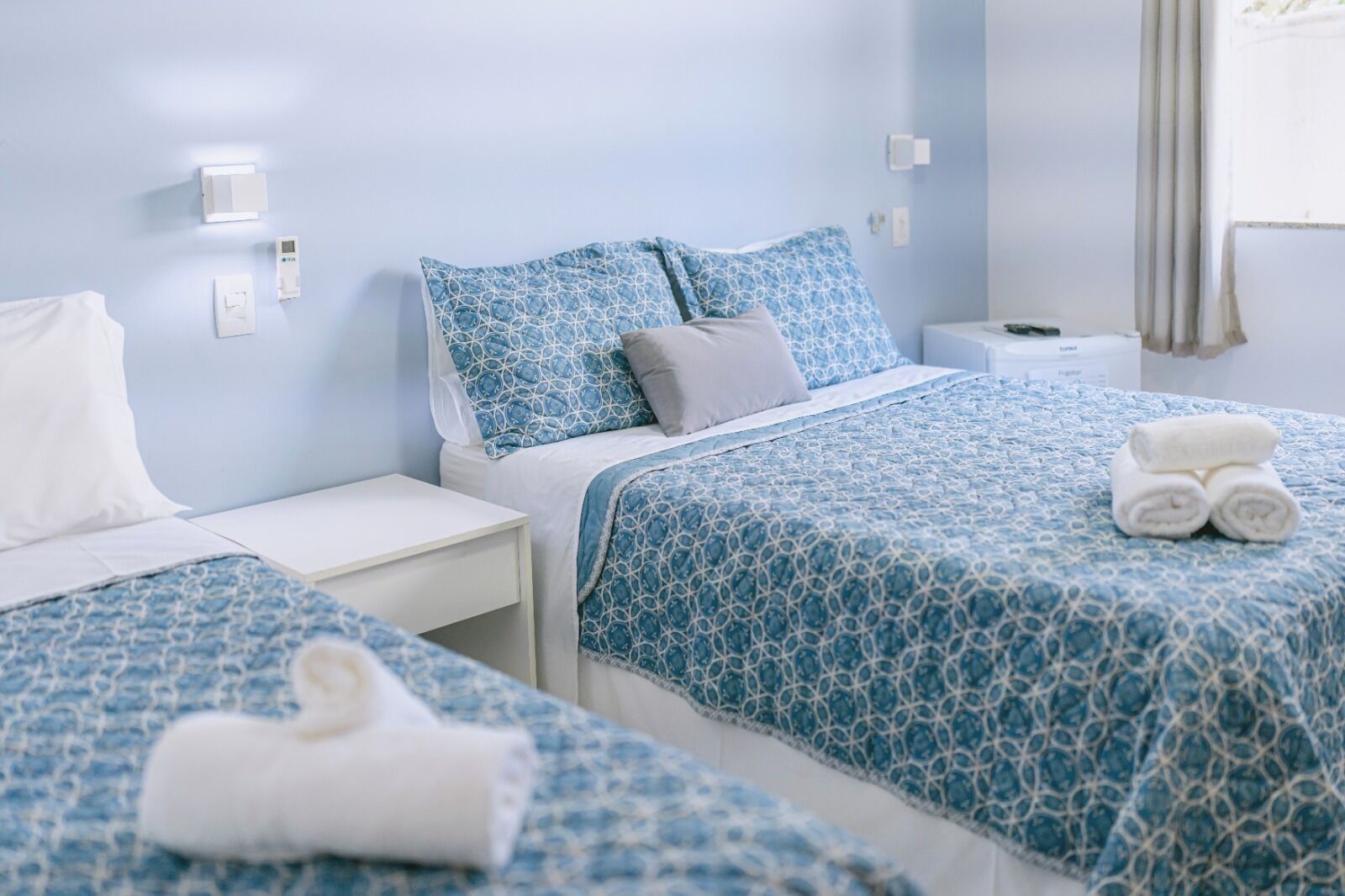 Camera d'albergo con due letti, ciascuno con una coperta e federe a motivi blu. Si nota un comodino bianco con una lampada, un piccolo tavolino da notte bianco e un asciugamano arrotolato sul letto.