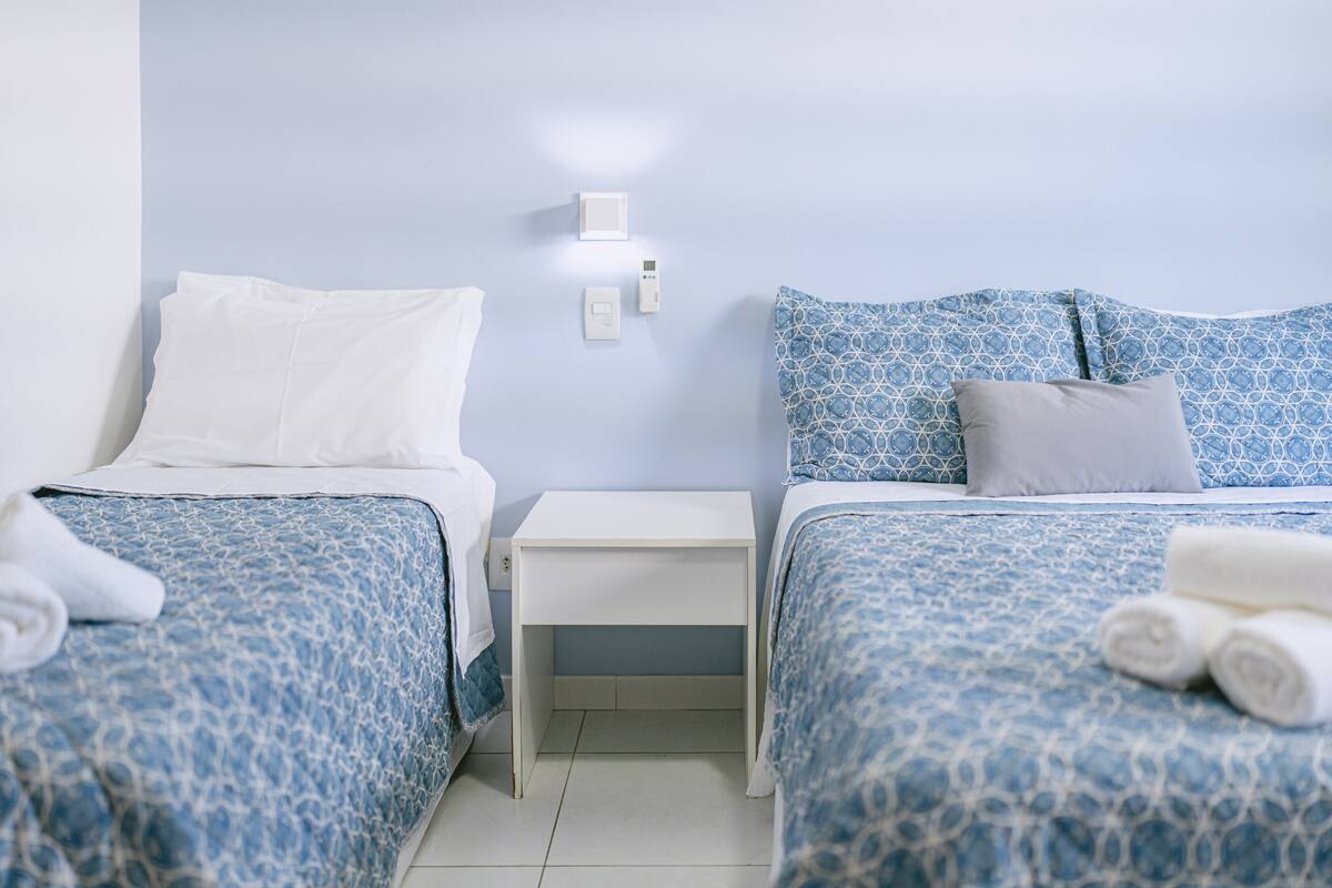 Ein Hotelzimmer mit zwei Betten, jeweils mit einer blauen Bettdecke und weißen Kissen. Dazwischen befindet sich ein weißer Nachttisch mit einer kleinen Schublade.