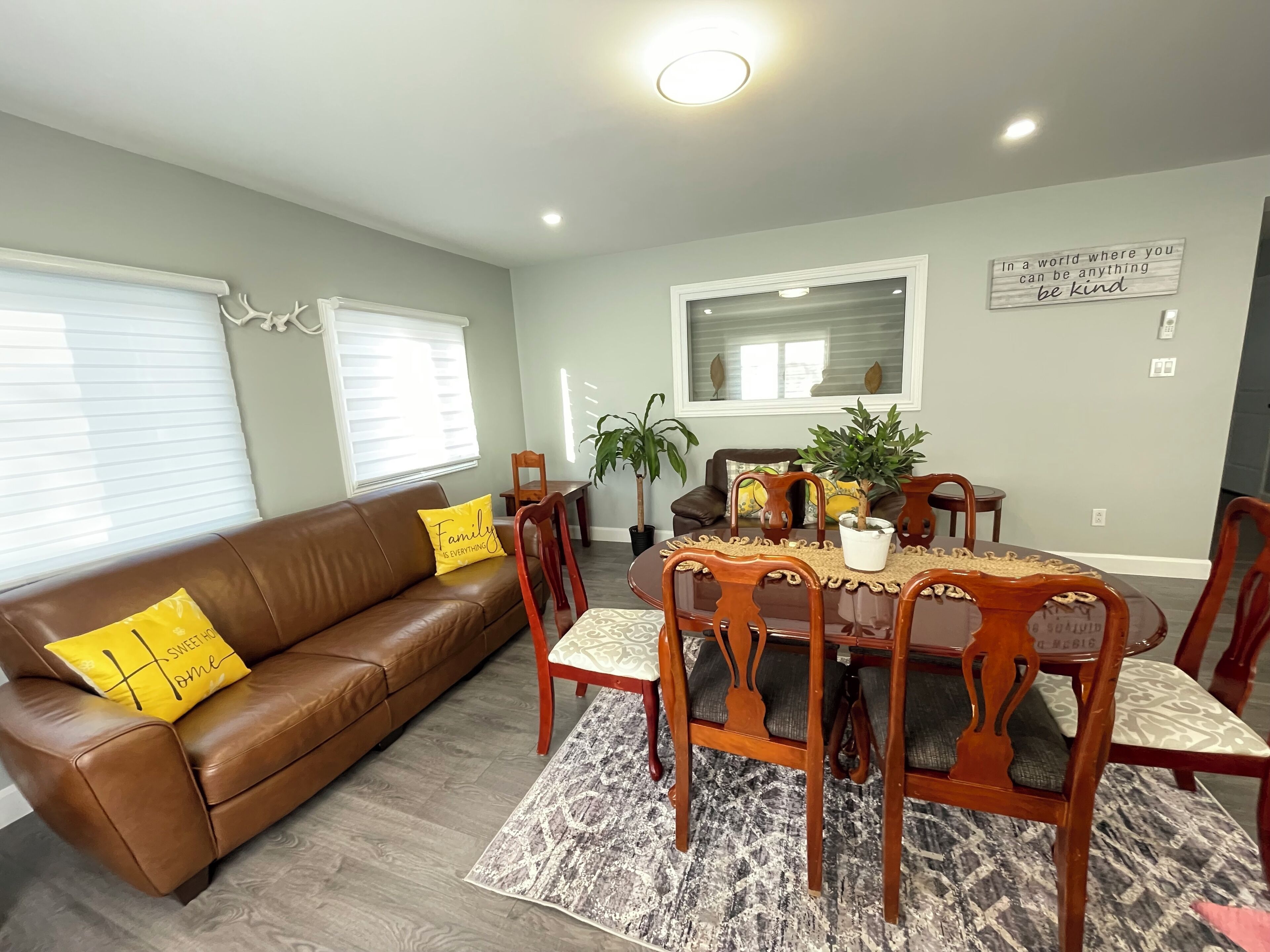 A dining area with a wooden table and chairs, a brown leather couch with yellow pillows, and a potted plant on the table.